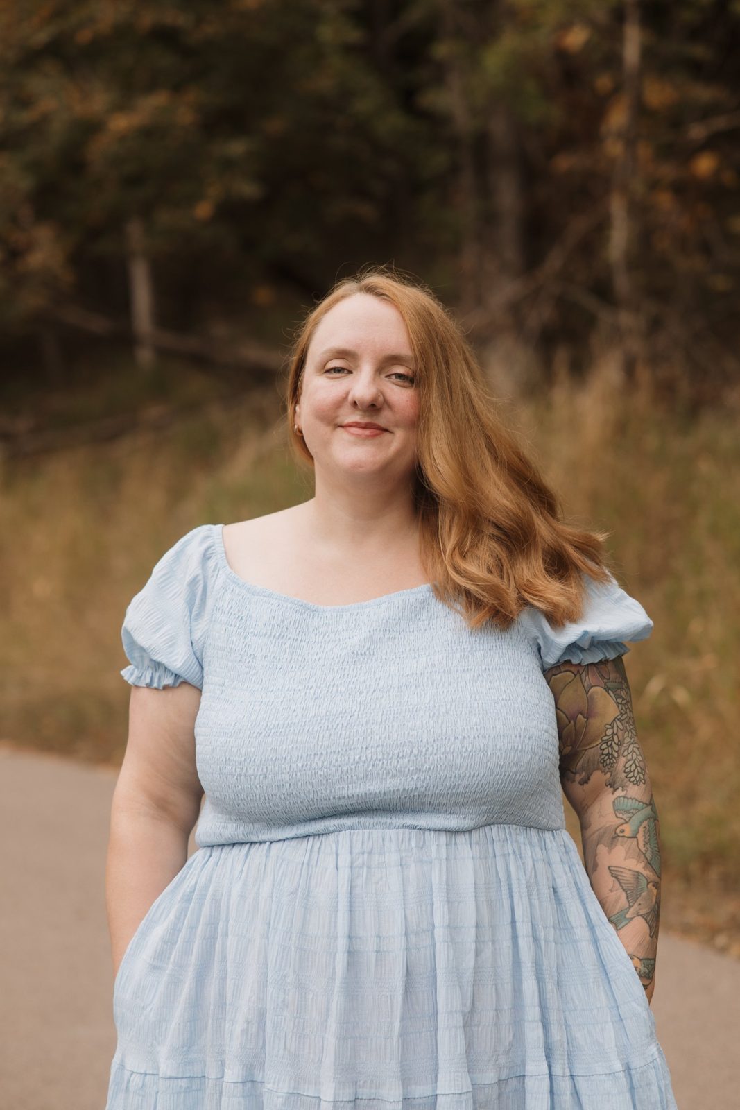 Hannah Kidder smiling, standing on a path in an autumn wood, wearing a light blue dress