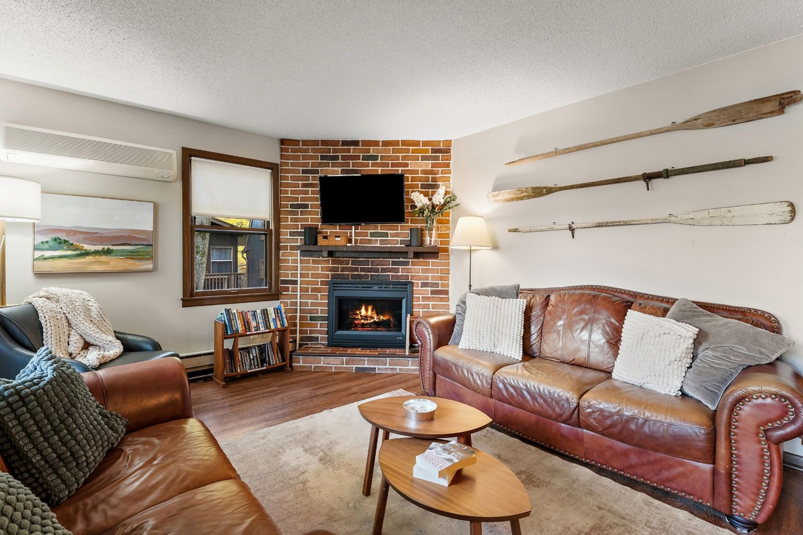 Living room with a lit brick fireplace, leather couches, chunky knit blankets, and oars hanging on the wall