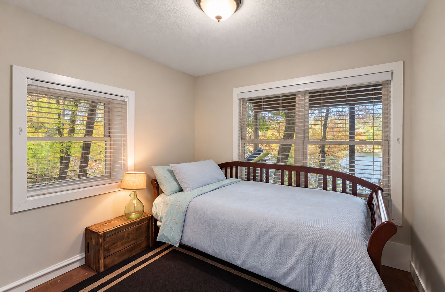 Bedroom with a wooden daybed in pale blue linens beside windows full of autumn trees
