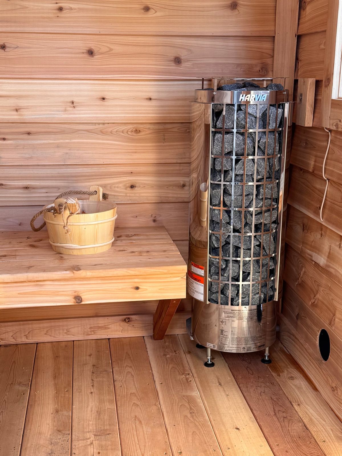 Inside a cedar sauna: a wooden bench, a water bucket with a ladle, and a stone-filled electric heater