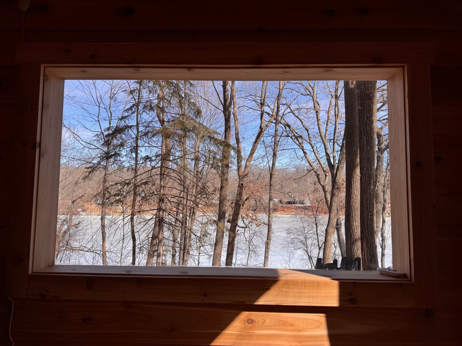 Looking out a cedar-framed window at bare trees and a frozen, snow-covered lake under a blue sky